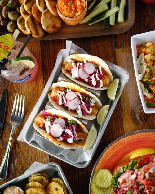 Three fish tacos topped with cabbage, radishes, and sauce, served with lime wedges, alongside various side dishes.