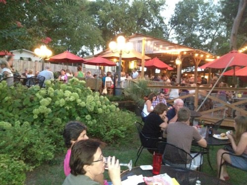 A lively outdoor restaurant scene with diners at tables under red umbrellas, surrounded by greenery and warm lighting.