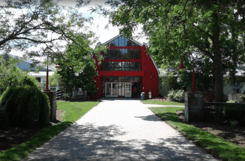A red building with large windows, surrounded by greenery and a paved pathway leading to the entrance.