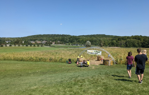 A sunny day at a corn maze entrance, with people walking towards it and a scenic green landscape in the background.