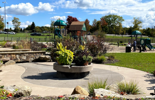 A park scene featuring a playground, landscaped gardens, and a clear blue sky with scattered clouds.