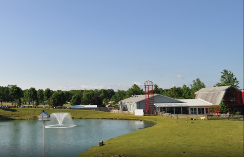A serene farm scene featuring a pond with a fountain, green grass, and a red barn under a clear blue sky.