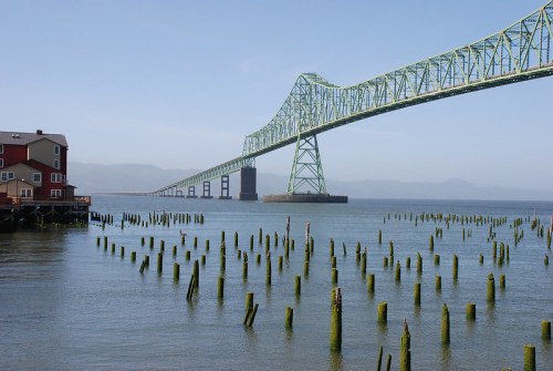 A green bridge spans over calm water, with wooden pilings visible and a red building on the shore in the foreground.