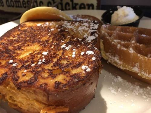 A close-up of a plate featuring French toast, a waffle, and a dollop of whipped cream, dusted with powdered sugar.