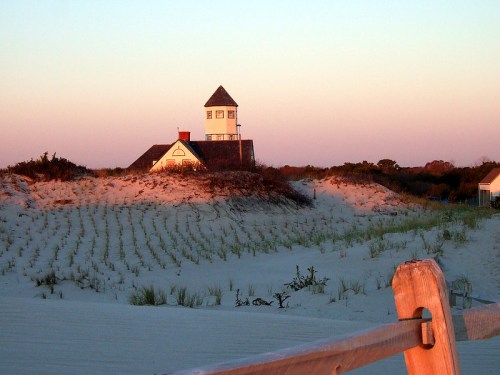 A lighthouse stands on a sandy dune at sunset, surrounded by grass and a wooden fence in the foreground.