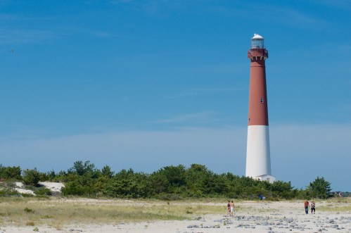 A tall lighthouse stands against a clear blue sky, surrounded by greenery and sandy beach, with people walking nearby.