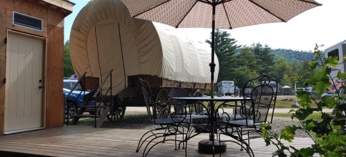 A wooden deck with a table and chairs, overlooking a covered wagon and a scenic outdoor area.
