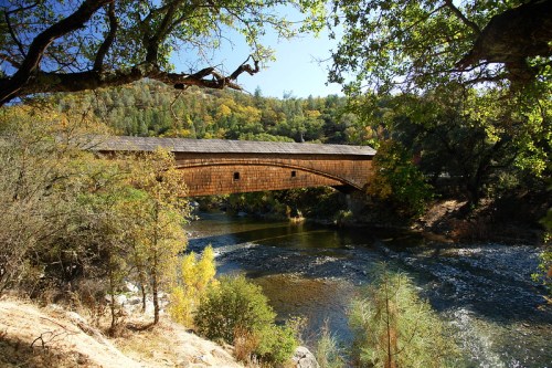 A wooden covered bridge spans a river, surrounded by trees and autumn foliage under a clear blue sky.