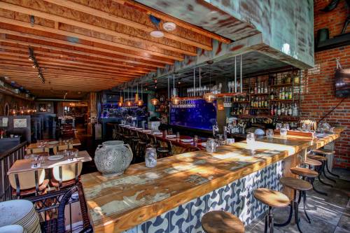 A stylish restaurant interior featuring a wooden bar, modern lighting, and a vibrant blue back wall with shelves of drinks.