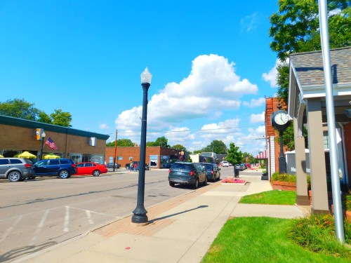 A sunny street scene with cars parked along the road, shops on the left, and a lamppost in the foreground.