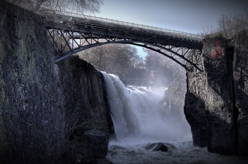 A waterfall cascades beneath a metal bridge, surrounded by rocky cliffs and mist.