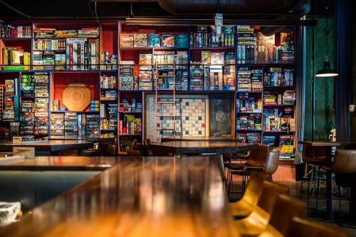 A cozy bar with a wooden counter and a wall filled with board games and a scrabble board in a vibrant setting.