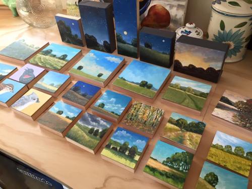 A collection of small landscape paintings displayed on a wooden surface, featuring trees, fields, and various skies.