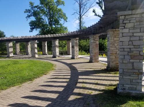 A stone pavilion with curved columns and shadows, surrounded by greenery under a clear blue sky.