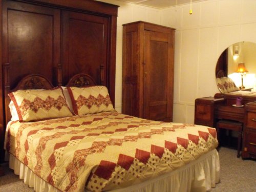 Cozy bedroom featuring a wooden bed with a quilt, a wardrobe, and a dresser with a round mirror. Warm lighting adds charm.
