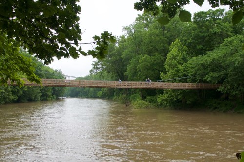 A wooden suspension bridge spans a muddy river, surrounded by lush green trees on a cloudy day.
