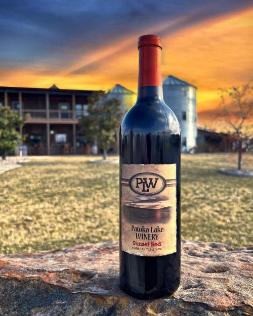 A bottle of Patoka Lake Winery Sunset Red wine against a vibrant sunset and a rustic building in the background.