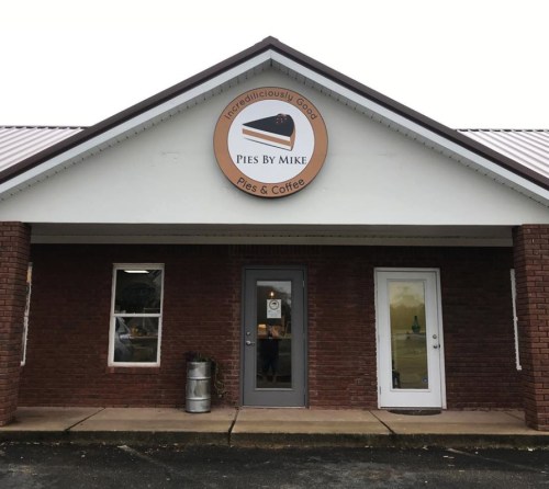 Exterior of a brick building with a sign reading "Pies By Mike" and two doors, one gray and one white.