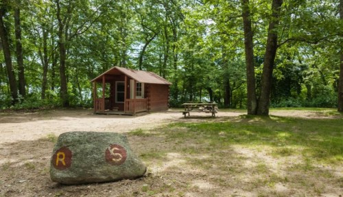 A small wooden cabin surrounded by trees, with a picnic table nearby and a large rock marked with "R" and "S."