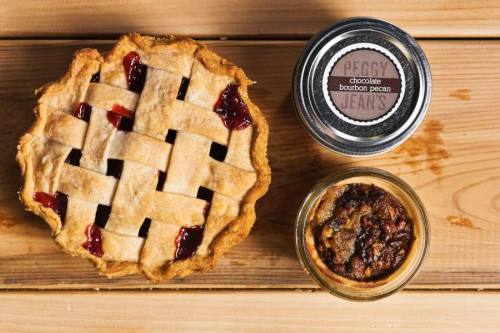 A lattice-top cherry pie next to a jar of chocolate bourbon pecan pie on a wooden surface.