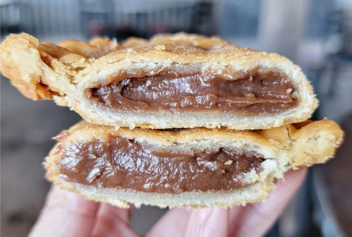 A hand holding a split pastry revealing a rich chocolate filling inside a golden, flaky crust.