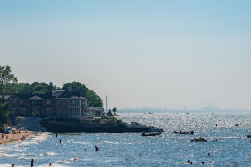 A sunny beach scene with people swimming, boats on the water, and a building near the shore under a clear blue sky.