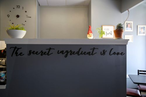 A wall with the phrase "The secret ingredient is love" in cursive, decorated with plants and framed photos.