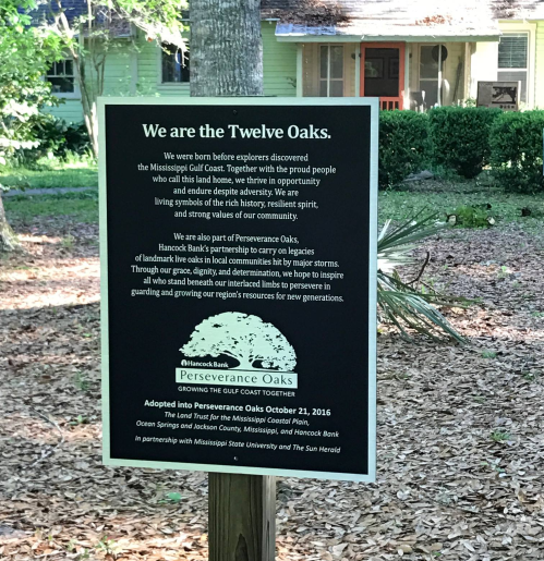 Sign at Twelve Oaks highlighting community heritage and preservation efforts, with a green house in the background.