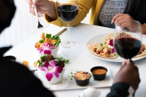 A table set with two glasses of red wine, a salad with flowers, and a plate of seafood, with hands reaching for food.