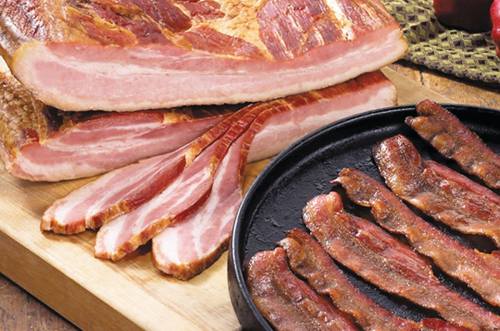 Sliced bacon and a frying pan with crispy bacon strips on a wooden surface.