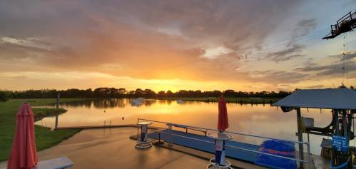 Sunset over a calm lake, with a dock and umbrellas in the foreground, surrounded by green fields and dramatic clouds.