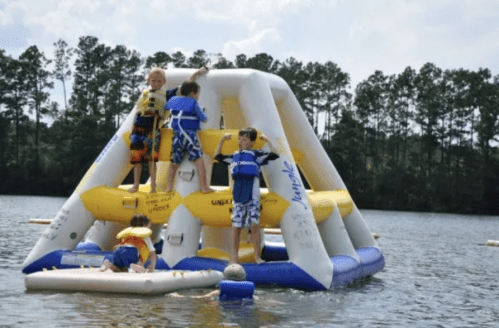 Children playing on an inflatable water structure in a lake, surrounded by trees and a sunny sky.