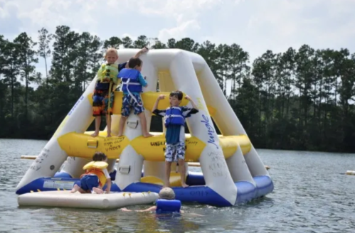 Children playing on an inflatable water structure in a lake, surrounded by trees and a sunny sky.