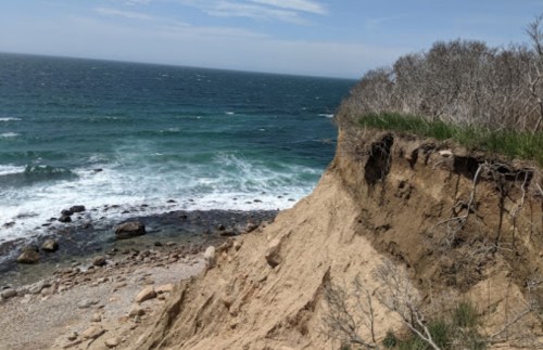 A sandy cliff overlooking a rocky beach and waves, with sparse vegetation and a clear blue sky above.