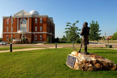 A bronze statue of a man stands on a rock in front of a brick government building with trees and a clear blue sky.