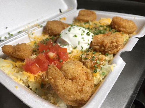 A takeout container filled with shrimp, cheese, sour cream, diced tomatoes, and chives over a bed of rice.