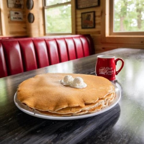A large stack of pancakes topped with two dollops of whipped cream, next to a red coffee mug on a table.