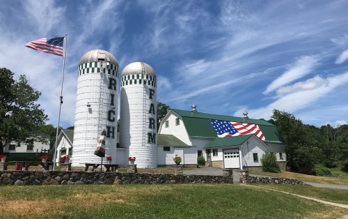 A farm with two silos, an American flag, and a barn featuring a large flag mural against a blue sky.