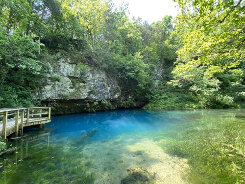 A serene blue pond surrounded by lush greenery and rocky cliffs, with a wooden platform at the edge.