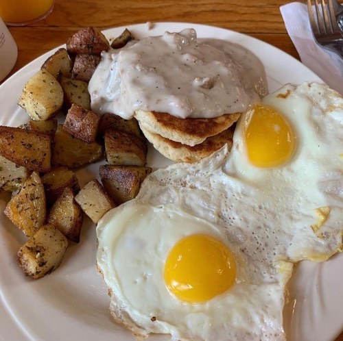 A plate of breakfast featuring two fried eggs, biscuits with gravy, and seasoned potato cubes.