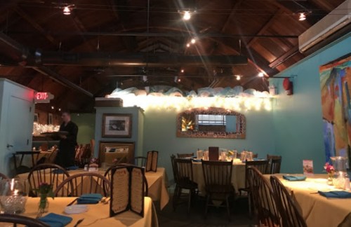 Cozy restaurant interior with wooden beams, blue walls, and tables set with yellow tablecloths and blue napkins.