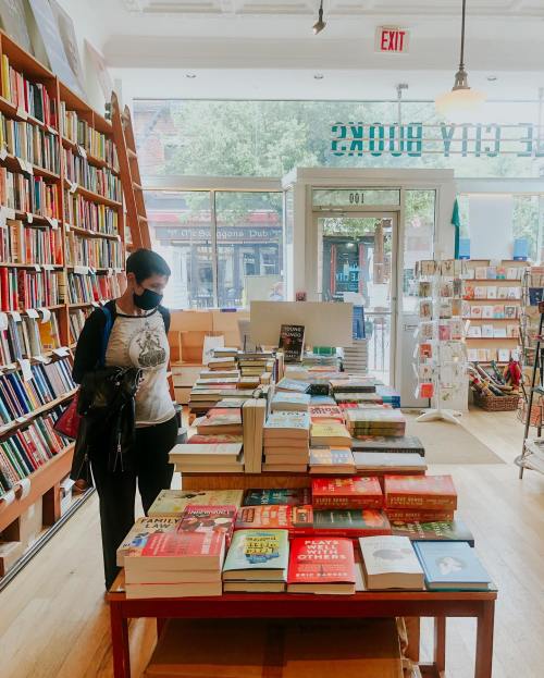 A person in a mask browses a table of books in a cozy bookstore with shelves filled with more books in the background.