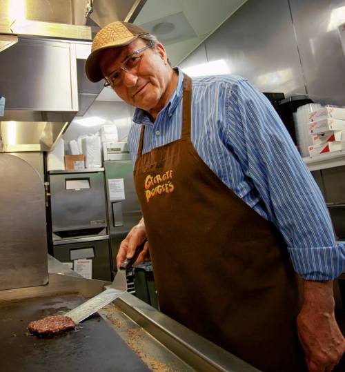 A smiling man in a brown apron stands at a grill, flipping a burger in a busy kitchen.