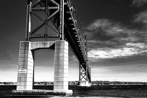 A black and white image of a large bridge spanning over water, with a clear sky and distant land in the background.