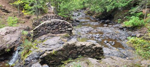 A rocky stream flows through a forested area, featuring a small stone bridge and lush greenery surrounding the water.