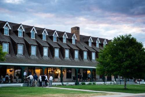 A historic hotel with a peaked roof, surrounded by green grass and people socializing on the porch under a cloudy sky.