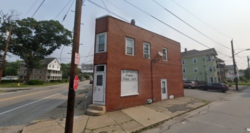 A two-story brick building on a corner lot, with a sign for Hartley's and nearby residential homes.