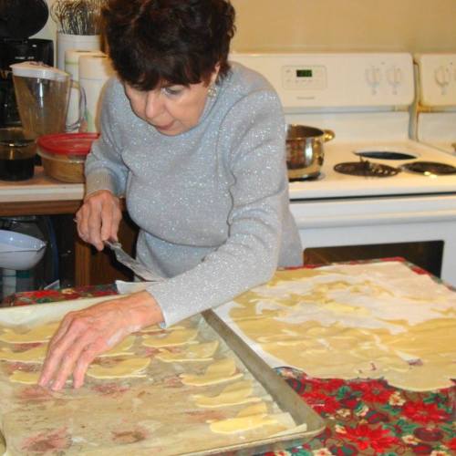 An older woman in a sparkly sweater prepares dough on a kitchen counter, focused on cutting shapes for cooking.
