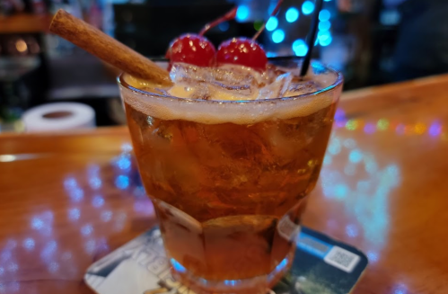 A close-up of a cocktail in a glass, garnished with cherries and a cinnamon stick, on a bar counter with festive lights.