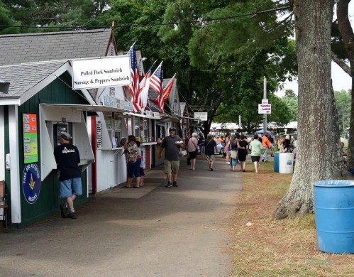 A bustling fairground with food stalls, American flags, and visitors walking along a tree-lined path.